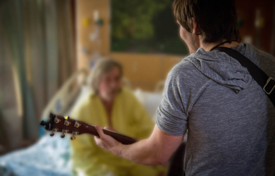 a musician with a guitar is playing in a hospital room. in a blurred background, you can see an older patient sitting upright in a hospital bed and is very engaged with the music