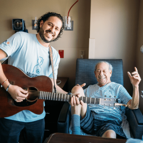 Volunteer Musician Bryant Del Toro poses with an older patient. Both are beaming with smiles after a wonderful Bedside performance